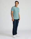 A man modeling a light blue crew-neck t-shirt, dark blue jeans, and white sneakers against a plain light gray studio background.