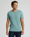 A man wearing a light teal short-sleeve crewneck t-shirt and dark blue jeans, standing against a solid light gray background.