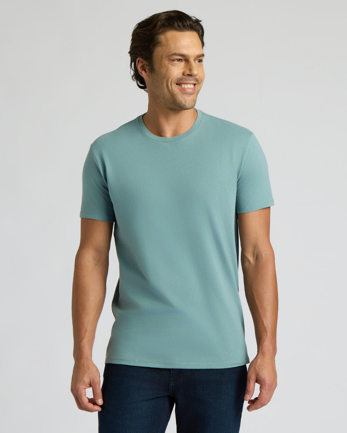 A man wearing a light teal short-sleeve crewneck t-shirt and dark blue jeans, standing against a solid light gray background.
