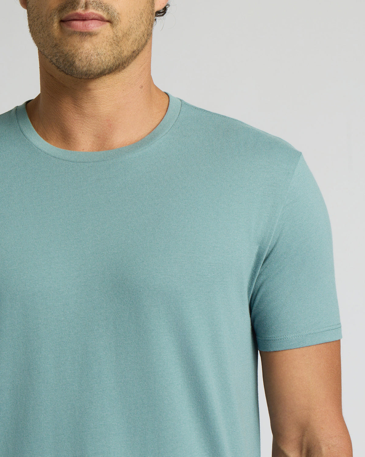 A close-up of a man wearing a short-sleeve crew-neck t-shirt in a light teal blue color, shown from the chin down against a neutral light gray background.