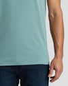 Close-up of a person wearing a light blue-green crew-neck t-shirt and dark blue jeans against a plain light gray background.