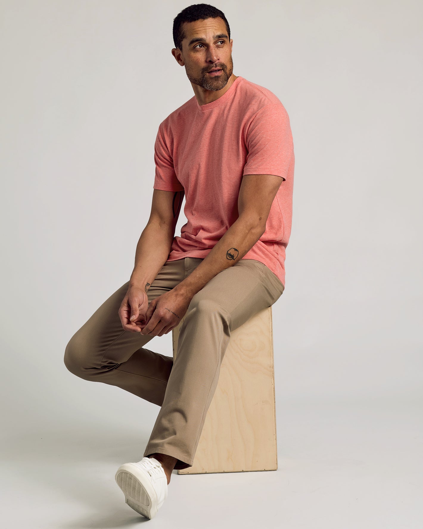 Man wearing Light Heather Vermilion Classic Crew Neck, seated on light-wooden cube.