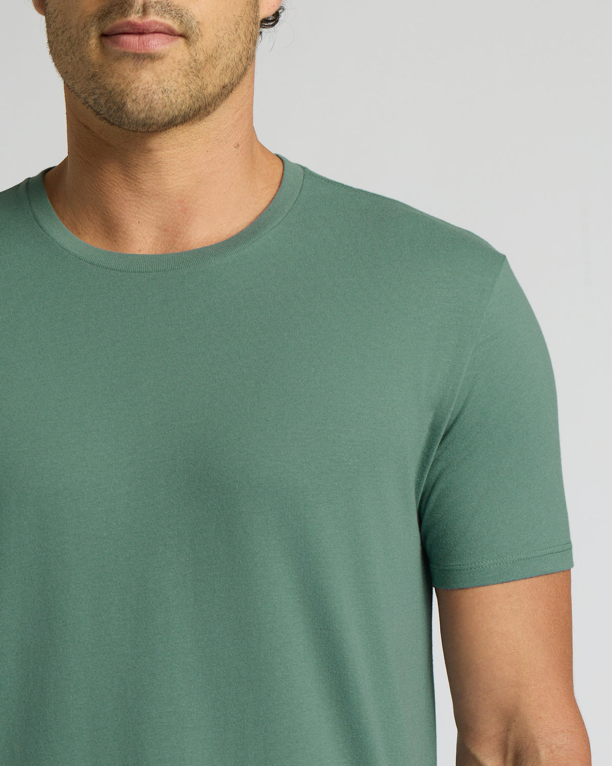 A close-up of a man wearing a muted sage green crew-neck t-shirt against a light gray background.