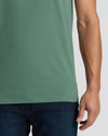 Close-up of a person wearing a muted sage green short-sleeve t-shirt and dark blue jeans against a light gray background.