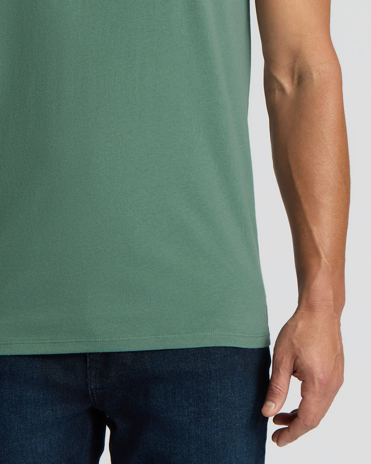 Close-up of a person wearing a muted sage green short-sleeve t-shirt and dark blue jeans against a light gray background.