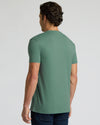 A man viewed from the back wearing a muted sage green short-sleeve t-shirt and dark blue jeans, standing against a light gray studio background.