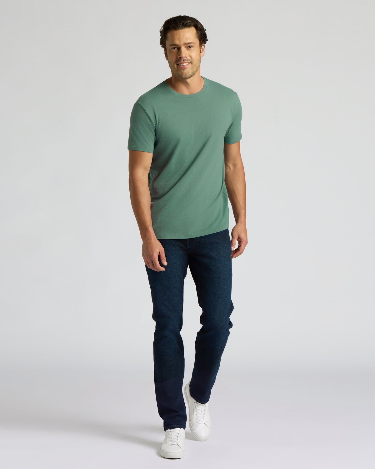 A man modeling a sage green crew-neck t-shirt, dark blue jeans, and white sneakers against a light gray studio background.