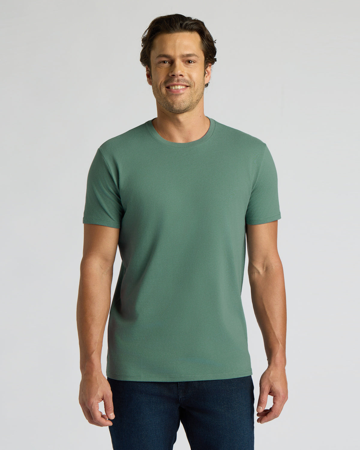 A man wearing a solid sage green crew-neck t-shirt and dark blue jeans, standing against a light gray studio background.