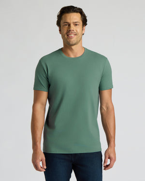 A man wearing a solid sage green crew-neck t-shirt and dark blue jeans, standing against a light gray studio background.