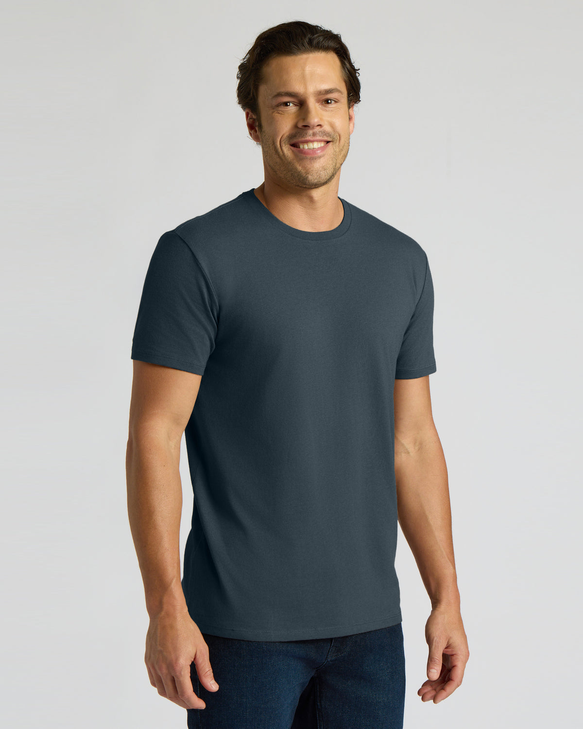 A man wearing a dark slate blue crewneck t-shirt and dark blue jeans, standing against a light gray studio background.