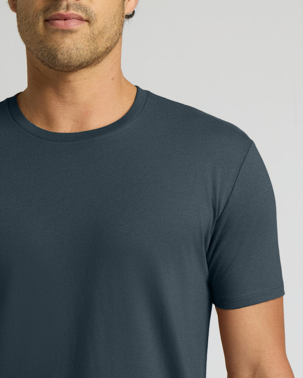 A close-up of a man wearing a dark slate blue crew-neck t-shirt against a light gray background.