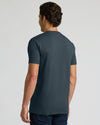 A man viewed from the back wearing a dark slate blue crewneck t-shirt and dark jeans, set against a plain light gray studio background.