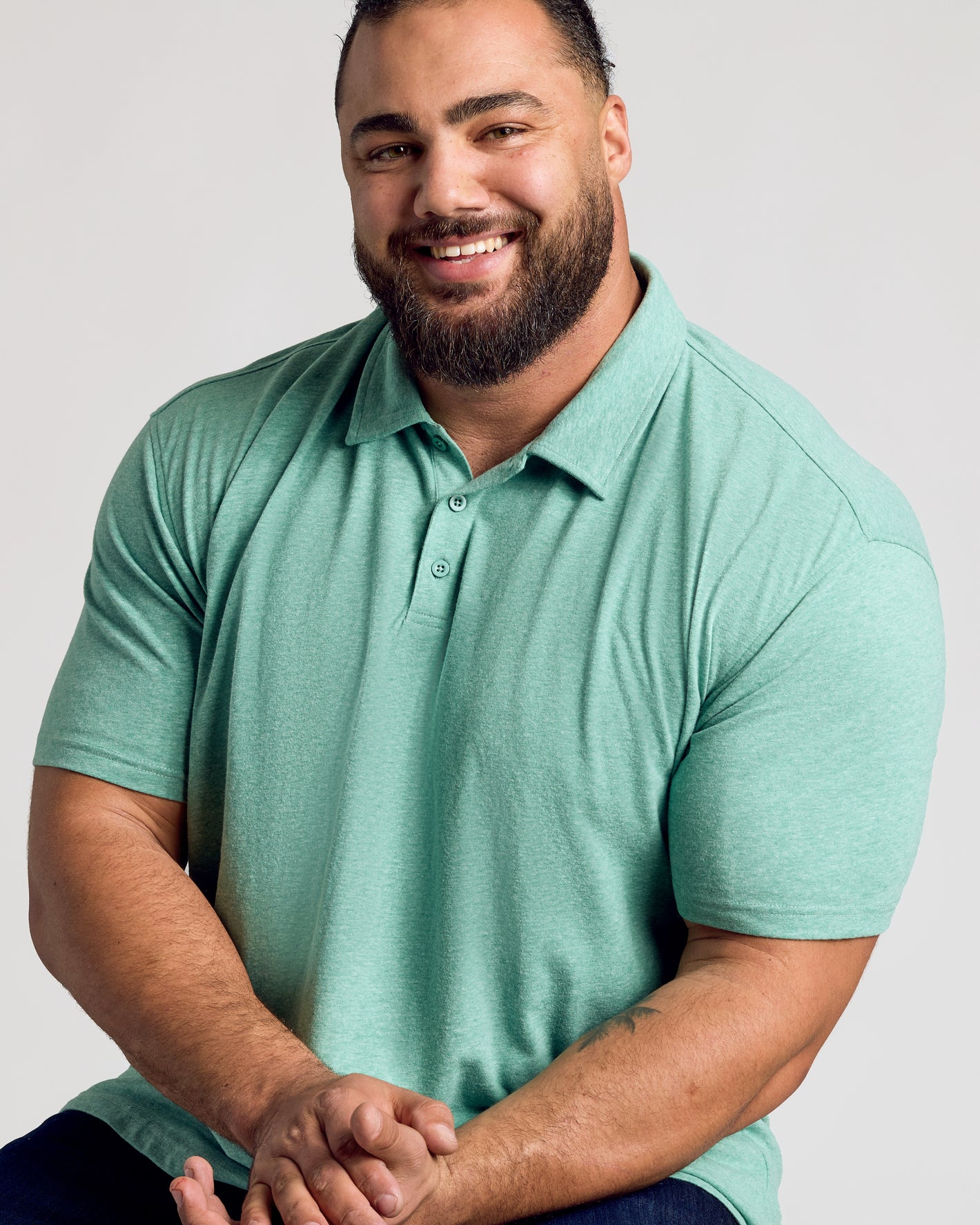 Man smiling, wearing Light Heather Greenery Classic Polo, seated with hands clasped.