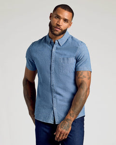 True ClassicA man wearing a light blue short-sleeve button-down shirt and dark blue jeans, standing against a light gray studio background.