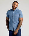 A man wearing a light blue short-sleeve button-down shirt and dark blue jeans, standing against a light gray studio background.