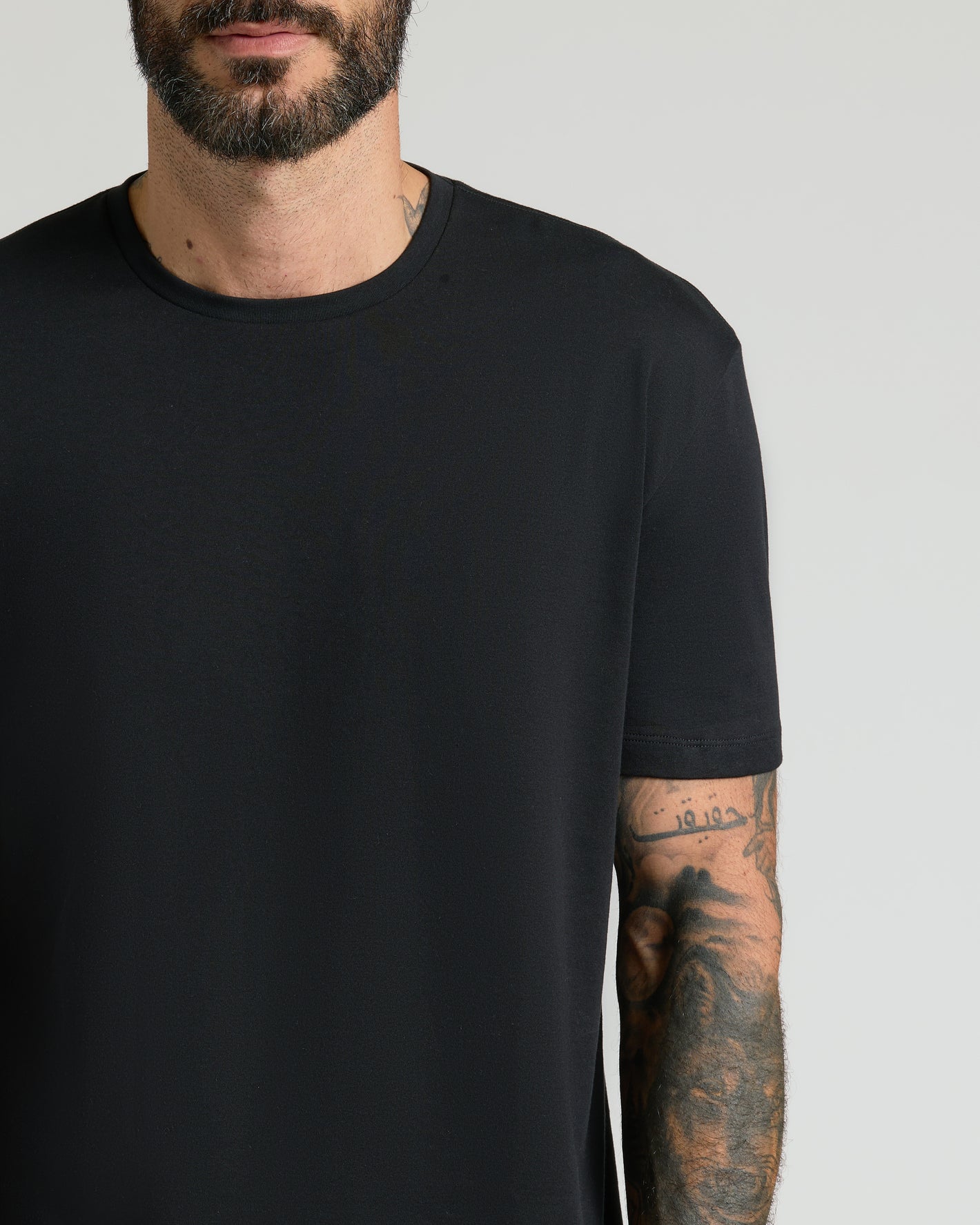 Black Premium Pima Crew Neck on man, tattooed forearm, neutral backdrop.