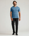 A man modeling a short-sleeve crewneck t-shirt in a dusty medium blue color, paired with black trousers and black shoes, standing against a light gray studio background.