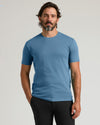 A man wearing a solid medium blue crew-neck t-shirt and black pants, standing against a light gray studio background.