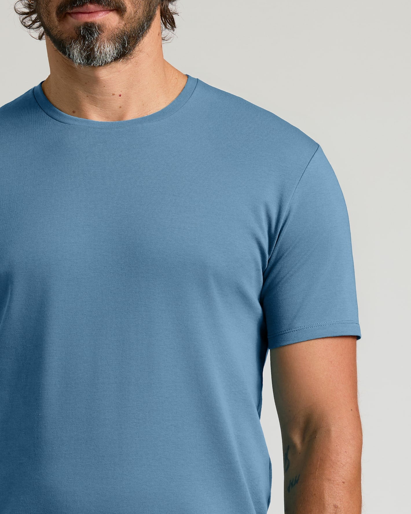 A close-up of a man wearing a solid medium blue crew-neck t-shirt against a light gray background.