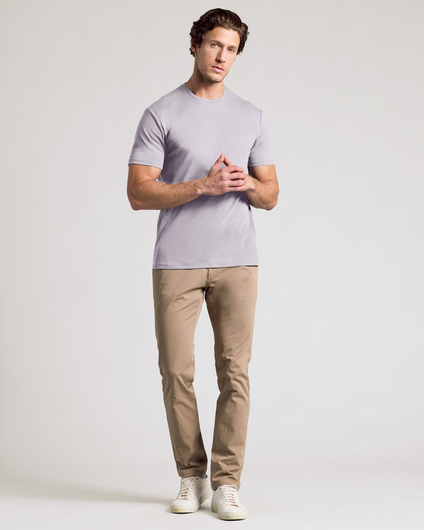 A man modeling a light lavender crew-neck t-shirt, khaki pants, and white sneakers against a plain light gray studio background.