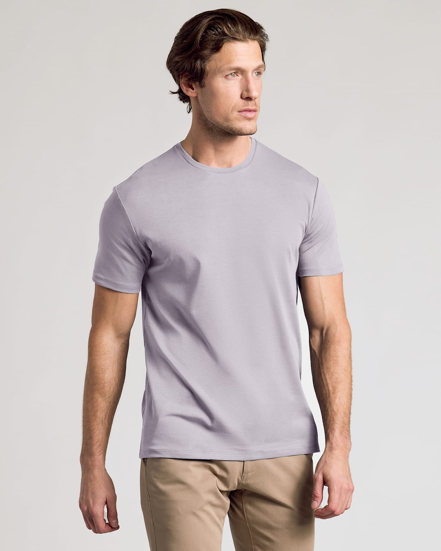 A man wearing a light lavender crew-neck t-shirt and khaki pants, standing against a neutral light gray background.