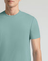 A close-up of a man wearing a short-sleeve crew-neck t-shirt in a muted sage green color, shown from the chin down against a light gray studio background.