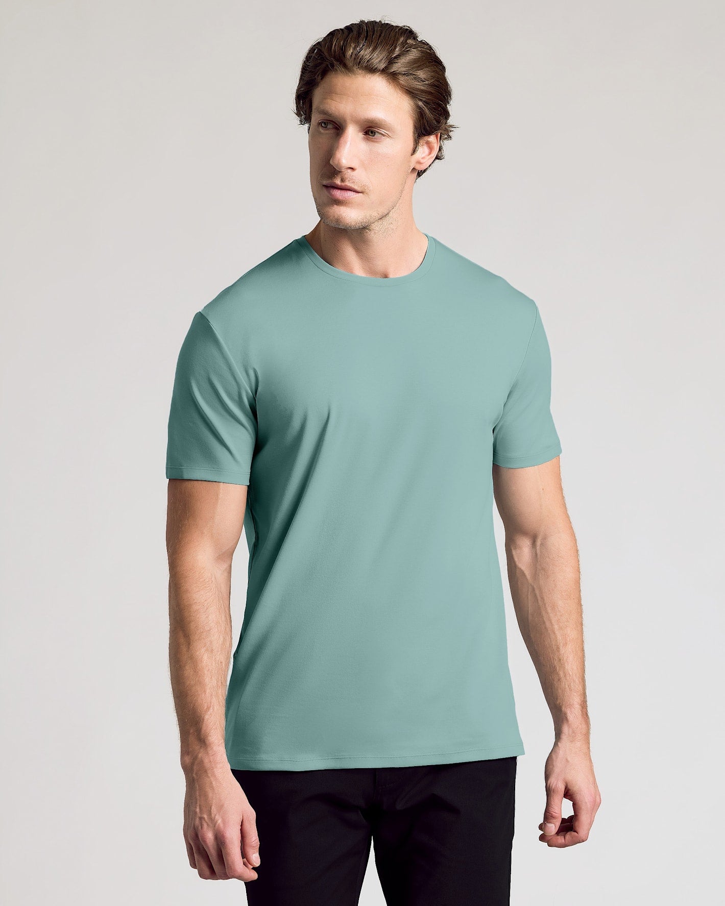 A man wearing a short-sleeve crewneck t-shirt in a muted sage green color, paired with black pants, standing against a light gray studio background.