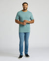 A man modeling a short-sleeve crewneck t-shirt in a dusty teal color, paired with blue jeans and black slip-on shoes, standing against a light gray studio background.