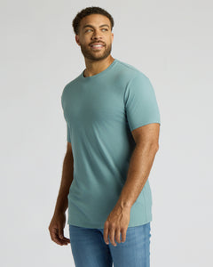 True ClassicA man wearing a fitted short-sleeve crewneck t-shirt in a muted teal-blue color, paired with blue jeans, standing against a light gray studio background.