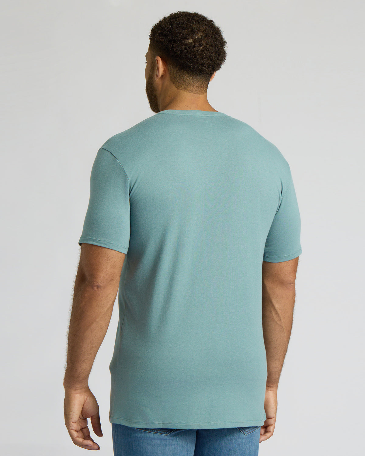 A man viewed from the back wearing a light blue-green crewneck t-shirt and blue jeans against a plain light gray studio background.