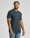 A man wearing a dark slate blue crewneck t-shirt and light blue jeans, standing against a light gray studio background.