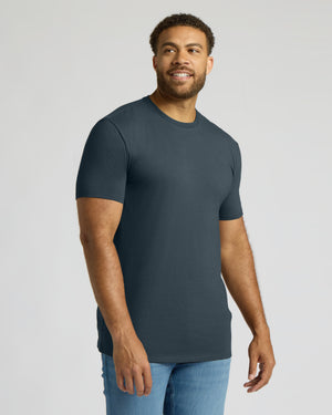 A man wearing a dark slate blue crewneck t-shirt and light blue jeans, standing against a light gray studio background.