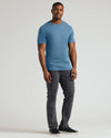 A man modeling a short-sleeve crewneck t-shirt in a dusty blue color, paired with dark gray slim-fit pants and black sneakers, against a light gray studio background.