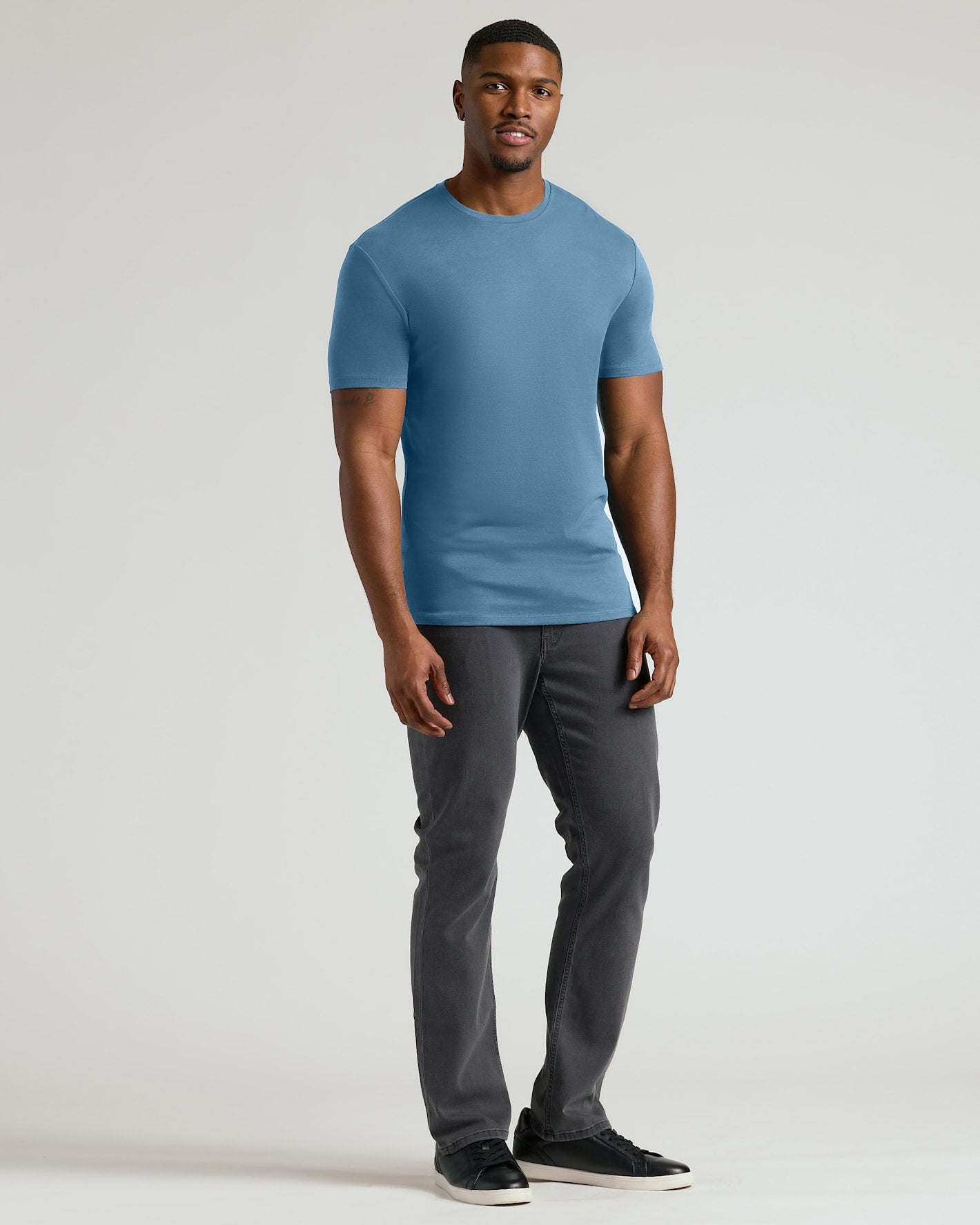 A man modeling a short-sleeve crewneck t-shirt in a dusty blue color, paired with dark gray slim-fit pants and black sneakers, against a light gray studio background.