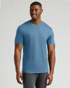 A man wearing a solid medium blue crewneck t-shirt and dark gray pants, standing against a light gray studio background.