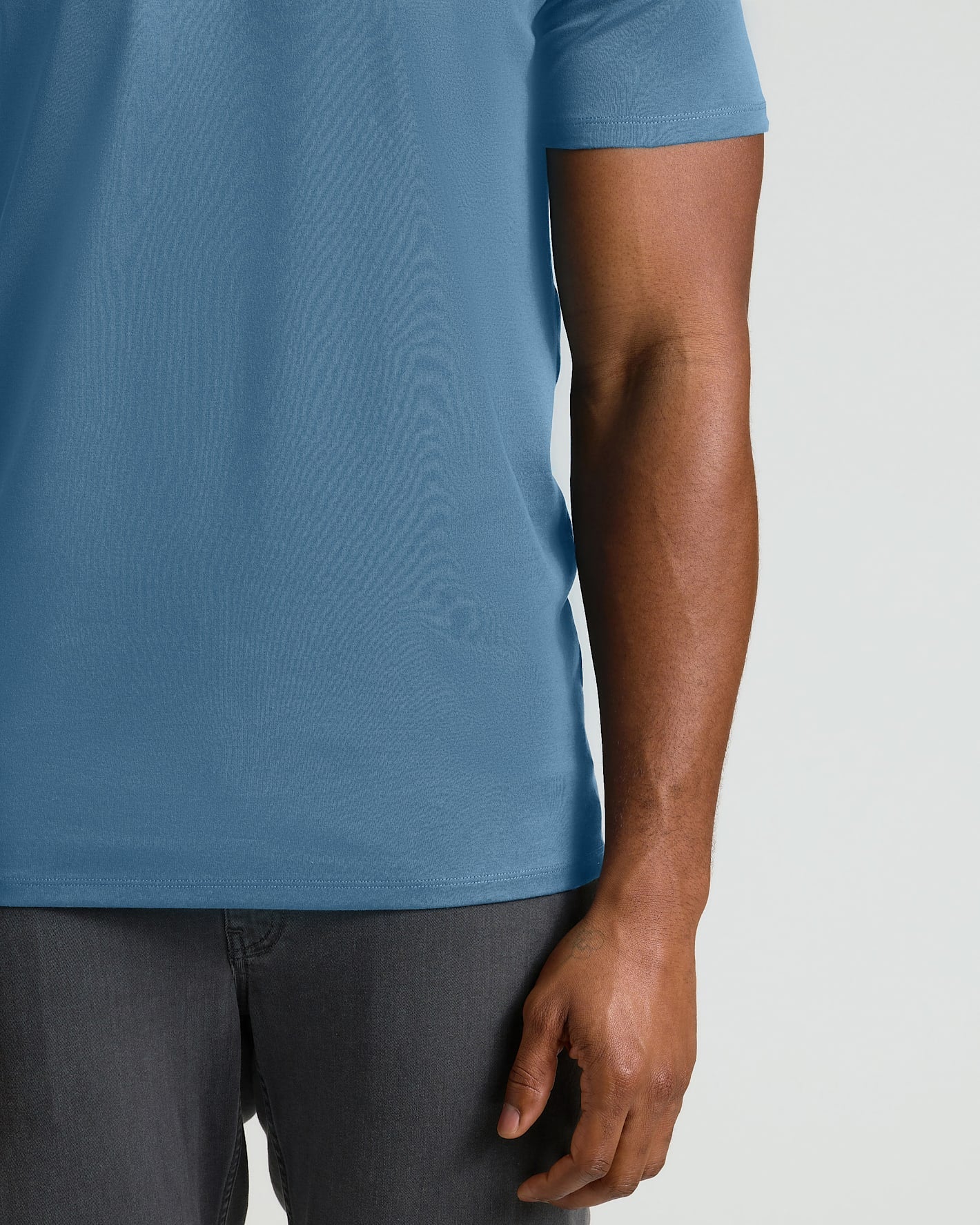 A close-up of a man wearing a short-sleeve crewneck t-shirt in a dusty blue color, paired with dark gray jeans, against a light gray background.