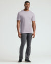 A man modeling a light purple crew-neck t-shirt and dark gray pants against a plain light gray studio background.