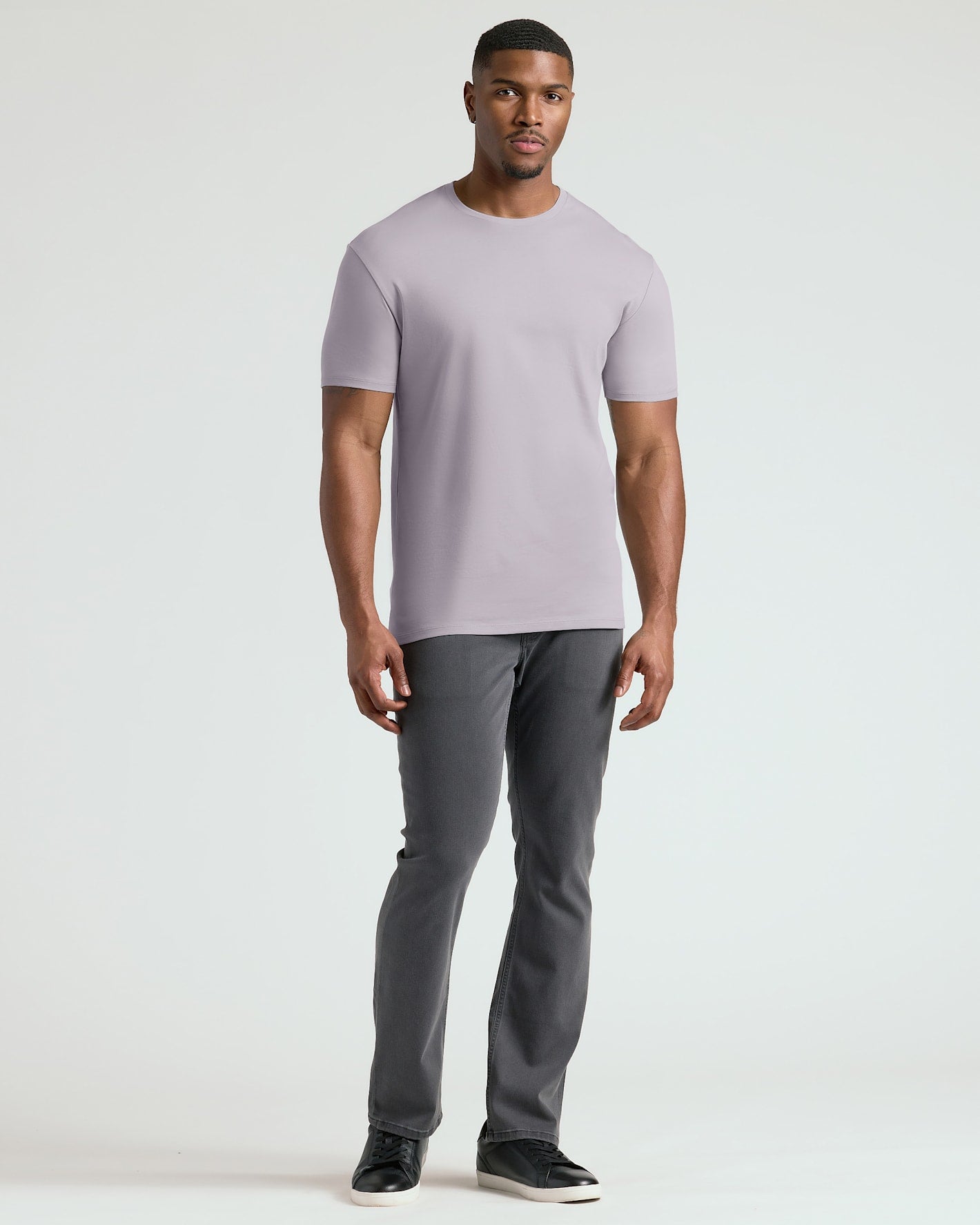 A man modeling a light purple crew-neck t-shirt and dark gray pants against a plain light gray studio background.