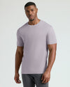 A man wearing a light lavender crew-neck t-shirt and dark gray pants, standing against a light gray studio background.