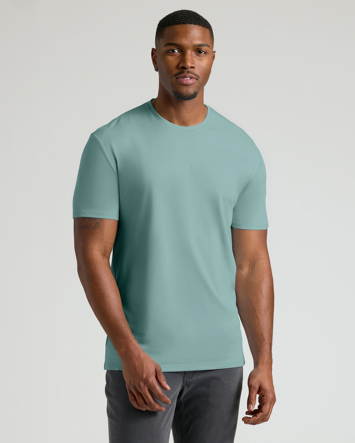 A man wearing a solid sage green crew-neck t-shirt and dark gray pants, standing against a light gray studio background.