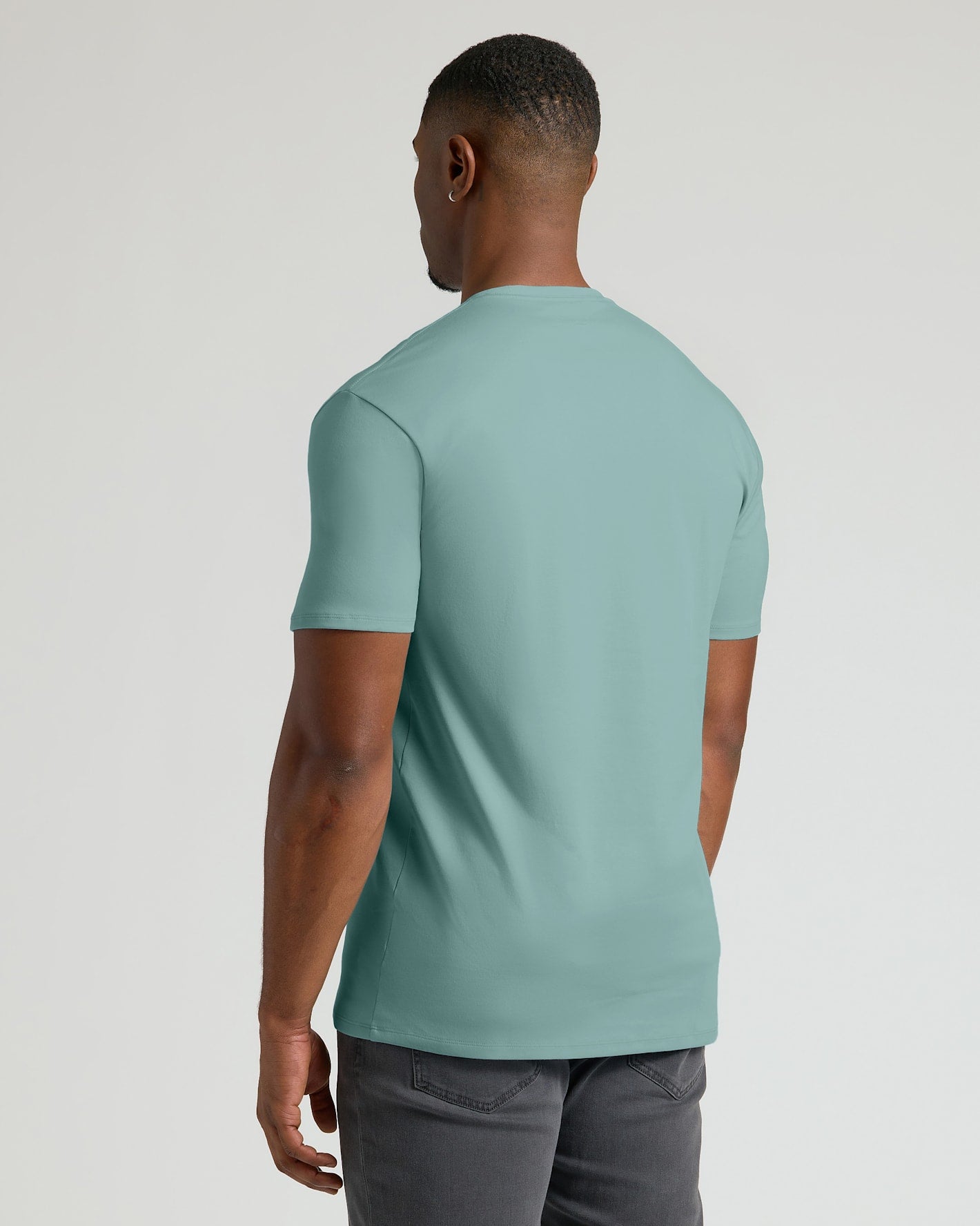 A man viewed from the back wearing a short-sleeve crew-neck t-shirt in a muted sage green color, paired with dark gray pants, against a light gray studio background.