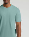 A close-up of a man wearing a solid sage green crew-neck t-shirt against a light gray studio background.