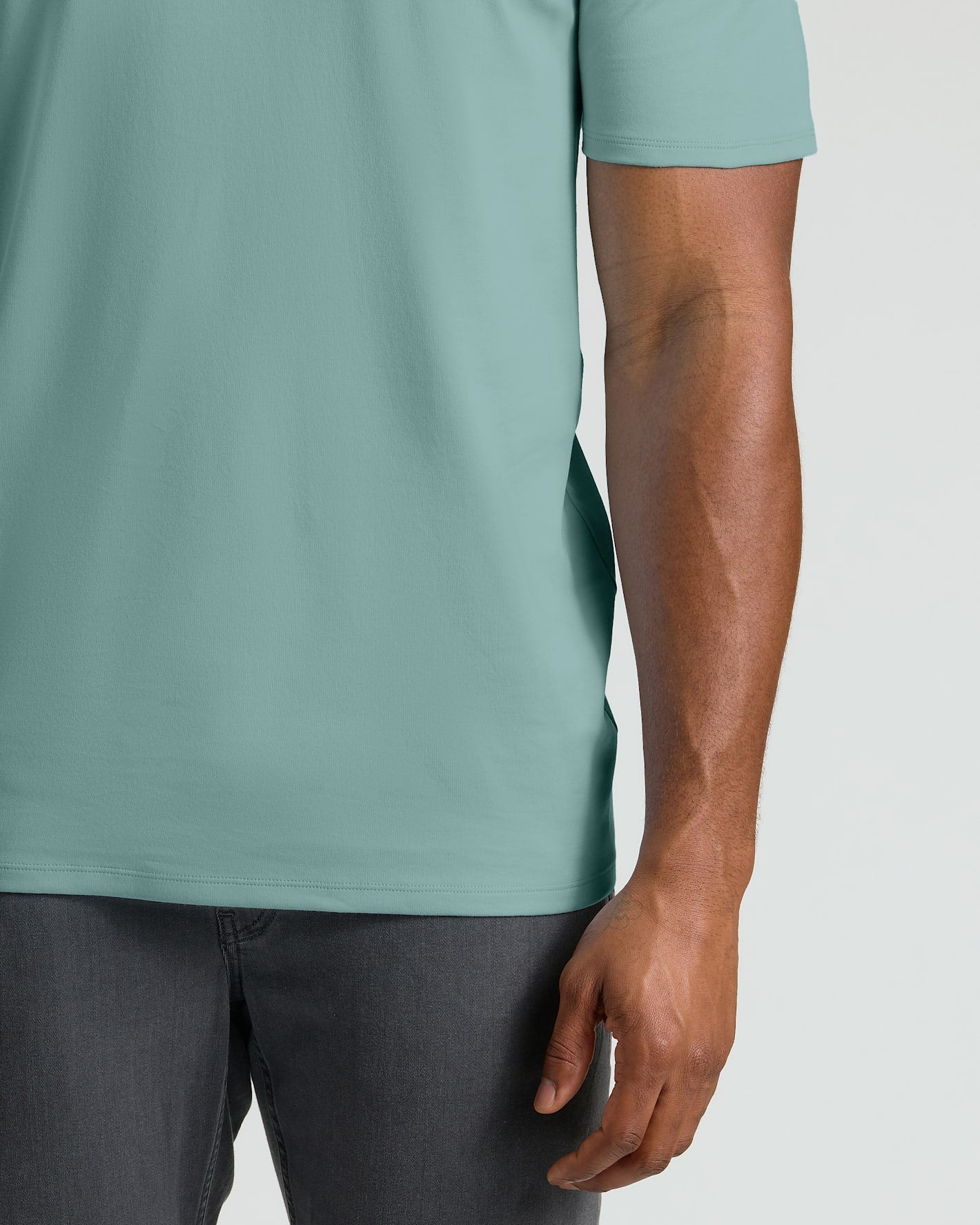 A close-up of a person wearing a short-sleeve crew-neck t-shirt in a muted sage green color, paired with dark gray jeans, against a light gray background.