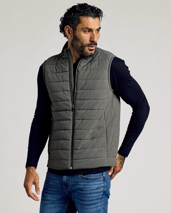 Man wearing a gray Carbon Puffer Vest over a black long-sleeve shirt and blue jeans, showcasing a casual and comfortable men's basics look.
