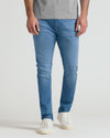 A person wearing slim-fit blue jeans, a heather gray t-shirt, and white sneakers, shown from the waist down against a light gray studio background.