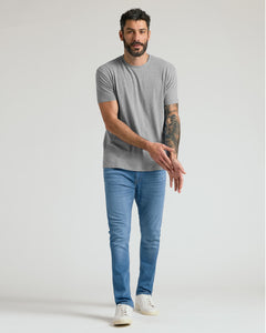 True ClassicA man modeling a light gray crew-neck t-shirt and blue slim-fit jeans with white sneakers, standing against a light gray studio background.