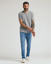 A man modeling a light gray crew-neck t-shirt and blue slim-fit jeans with white sneakers, standing against a light gray studio background.