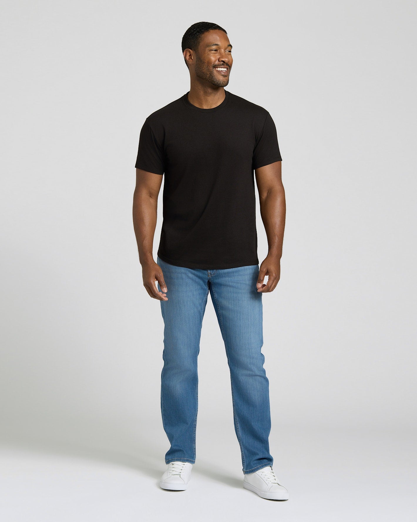 Man wearing a black t-shirt, blue jeans, and white sneakers on a plain background. MODEL_INFO: Model is 5'8" wearing size M short