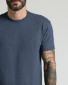 A close-up of a man wearing a short-sleeve crewneck t-shirt in heathered navy blue, showing his tattooed arm against a light gray background.