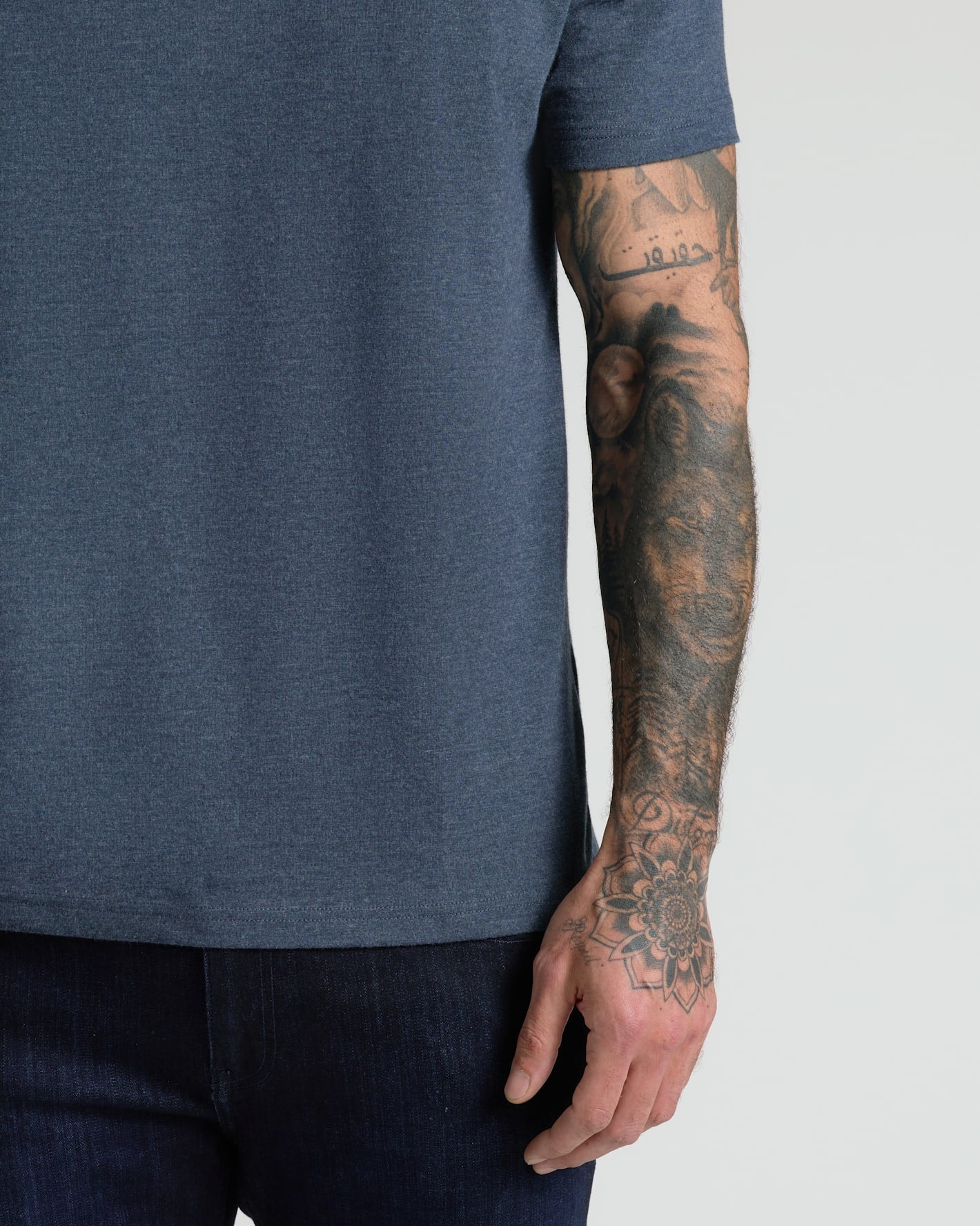 A close-up of a man's tattooed arm and hand wearing a dark heather blue short-sleeve t-shirt and dark jeans against a light gray background.